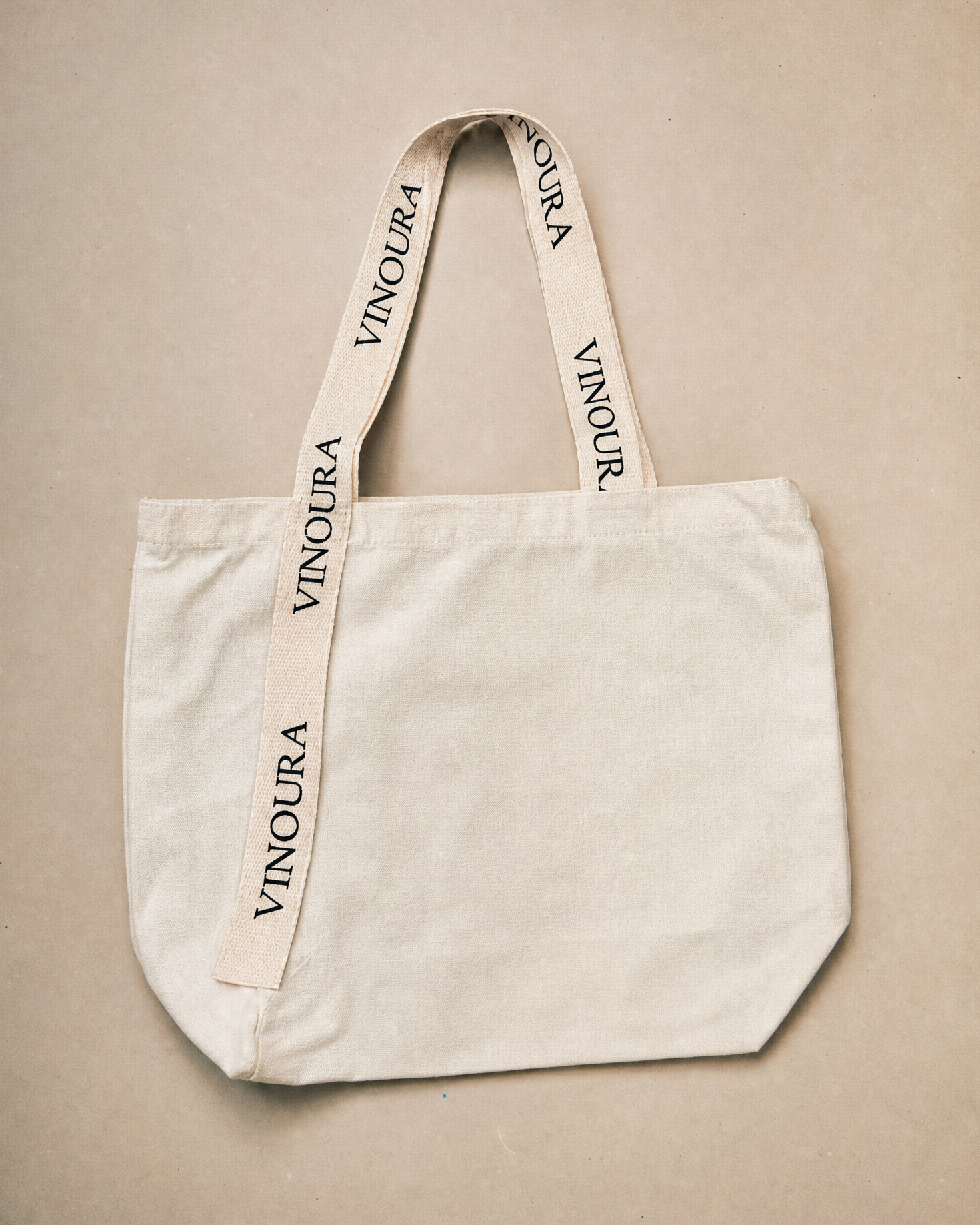 An off-white canvas tote bag with wide shoulder straps printed with the brand name 'VINOURA' in black, laid flat on a neutral beige background.