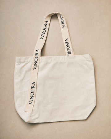 An off-white canvas tote bag with wide shoulder straps printed with the brand name 'VINOURA' in black, laid flat on a neutral beige background.