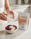 Person adding coconut flakes to a bowl of berries and chia seeds with a bag of vinoura shredded coconut in the background.